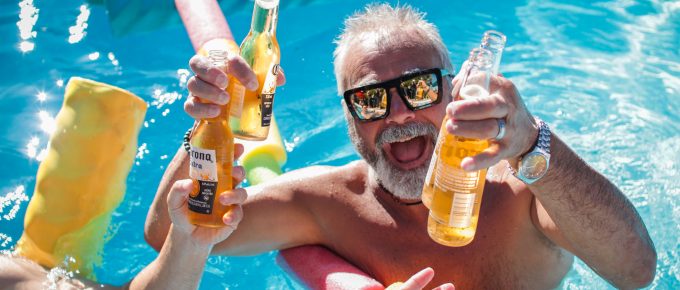 older man in a pool smiling with a beer in each hand